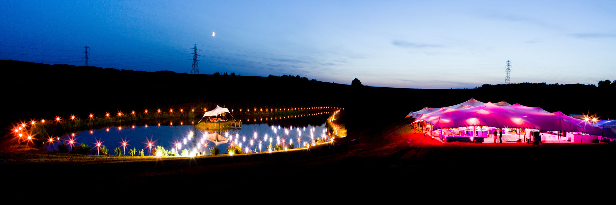 White stretch tent lit up at night, by a lake with lights around the lake.