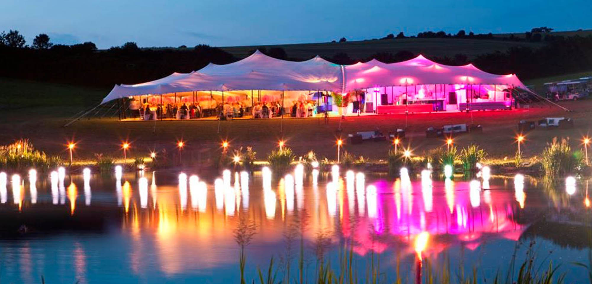 Header_Lighting_Lake_10x15 Stretch Tents at night by a lake lit up in colour LED lights