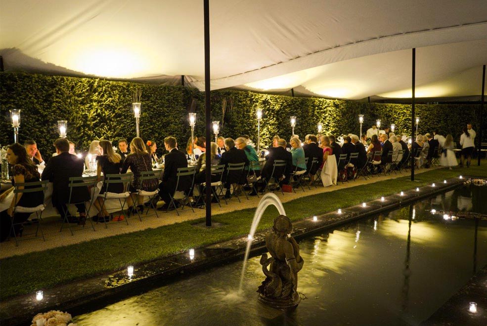 White stretch tent over long tables, hedges and ornamental pond at night