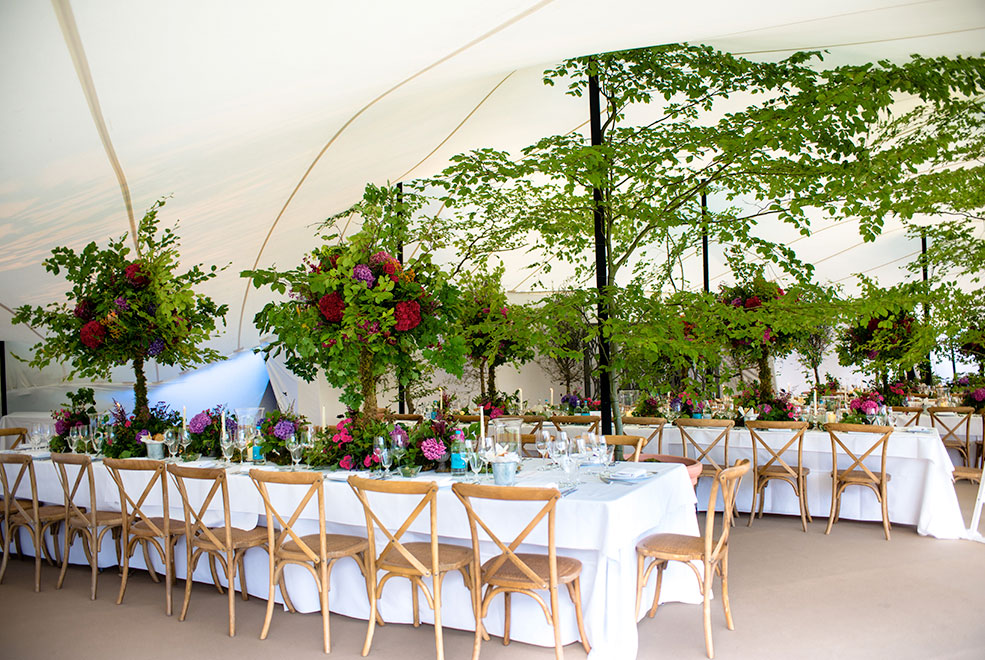 White Stretch tent with white long tables, set up with ink red flowers and huge green trees decorating the interior